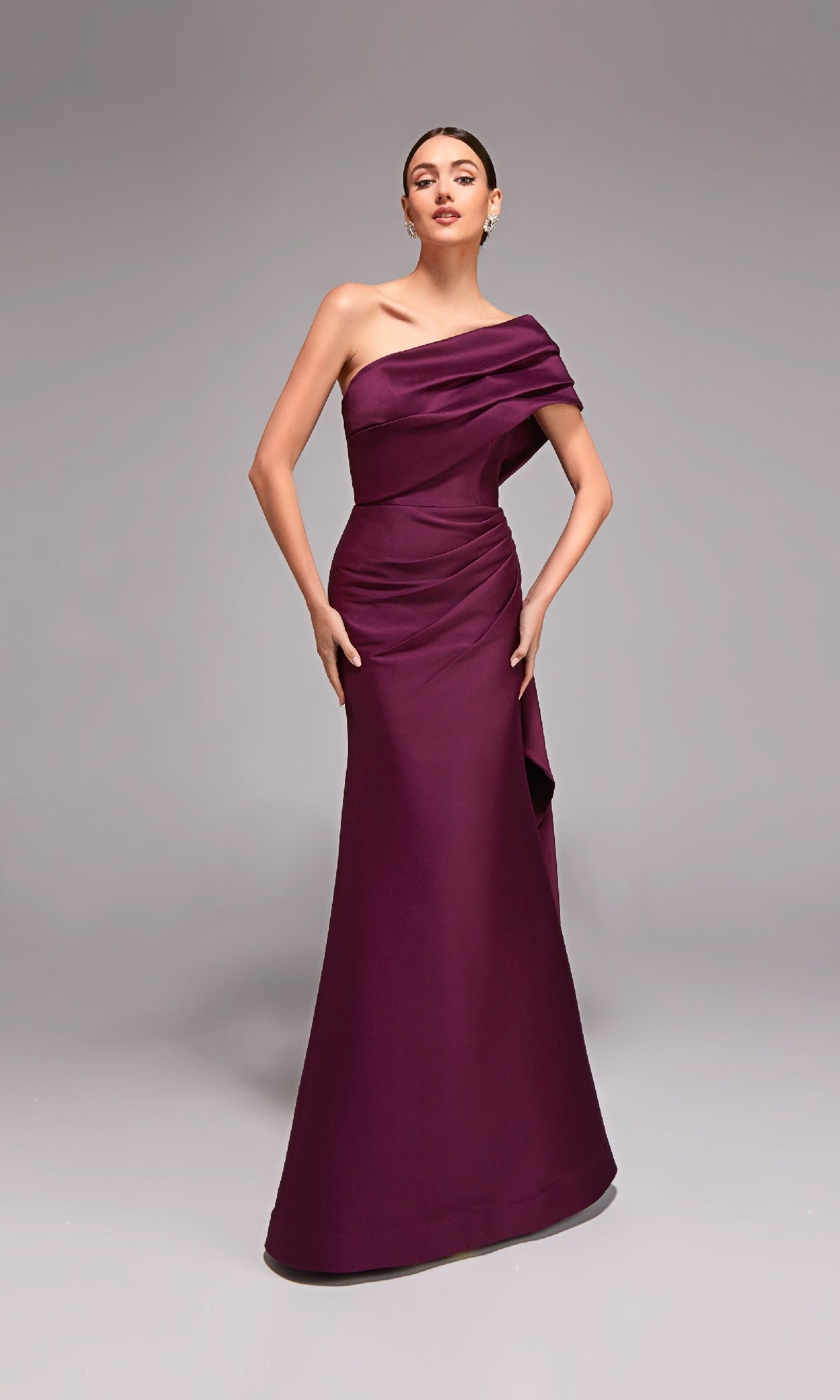 A woman models the Alyce 27838 long formal dress, featuring a one-shoulder design in dark purple, standing against a plain gray background.